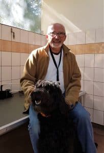 Bernard est assis dans le local détente de l'École de Chiens Guides de Paris, il sourit. Un labradoodle est assis entre ses jambes. 