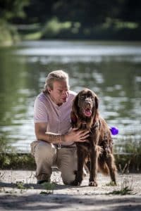 Olivier et assis aux côtés d'Orient qui est debout avec un gilet "chien guide en détente". Orient porte le bandana d'Olivier autour du cou, le binôme est devant un lac ensoleillé.