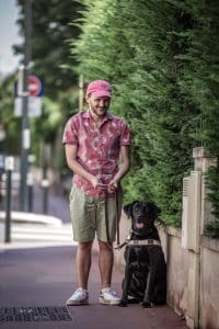 Geoffrey, souriant, est debout aux côtés de Pinaï, assise, qui porte son harnais de chien guide. 