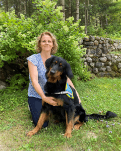 Frédérique Beaurain est assise dans l'herbe, elle a un hovawart avec un body élève chien guide à côté d'elle