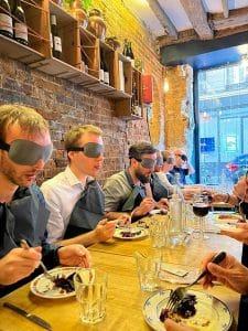 Plusieurs convives sont assis autour d'une table les yeux bandés, ils s'apprêtent à déguster leur plat. 