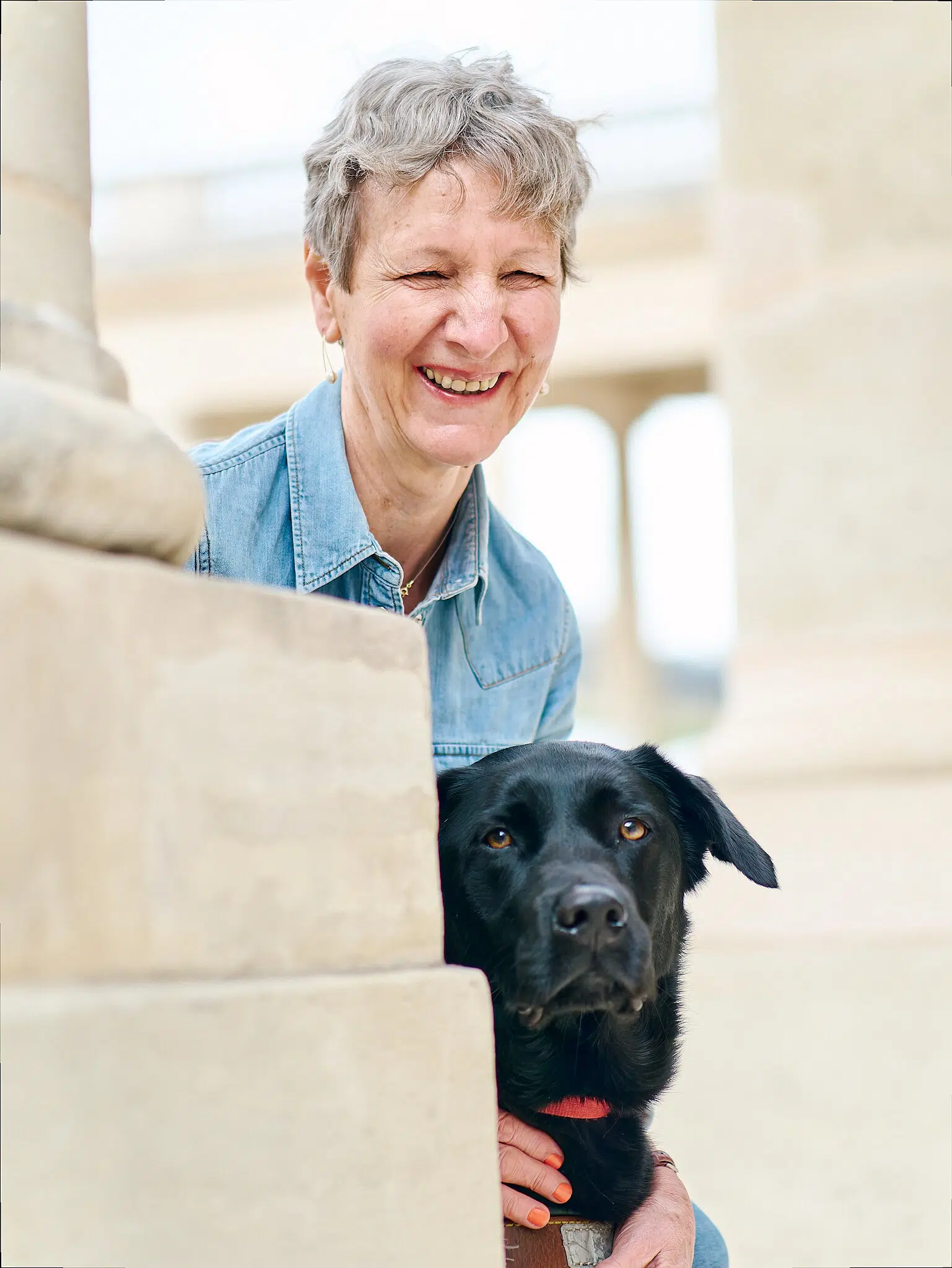 Martine, maîtresse de chien guide, avec Rusty, chien guide noir, qui se cachent derrière un mur et regardent l'objectif